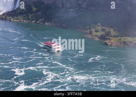 Una vista aerea cattura una barca turistica piena di visitatori con poncho rossi che naviga sulle vibranti acque turchesi alla base delle cascate del Niagara, Ontario, Foto Stock