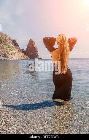 Donna spiaggia donna in vestito nero in piedi nel mare vicino a scogliere rocciose in una giornata di sole sistemandosi i capelli lunghi Foto Stock