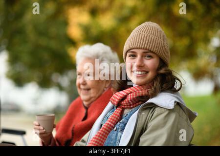 Ritratto di una nipote e di una nonna sedute sulla panchina nel parco autunnale, bevendo un caffè e ridendo insieme. Foto Stock