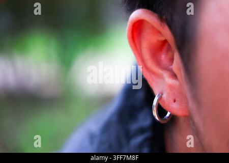 Un orecchio umano, orecchino a cerchio in argento, primo piano, messa a fuoco poco profonda, sfondo all'aperto, dettaglio pelle, gioiello, lobo dell'orecchio con piccolo neo, capelli scuri, verde sfocato N Foto Stock