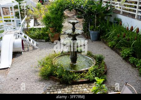 Un cortile giardino con fontana a più livelli, vegetazione rigogliosa, percorso di ghiaia con pianta in vaso, aiuola fiorita, elemento acquatico, recinto bianco, scivolo e spazio esterno tranquillo Foto Stock