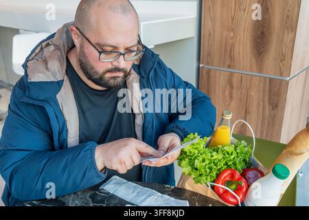 Uomo preoccupato controlla la ricevuta della spesa seduto in un supermercato con una borsa della spesa piena di prodotti alimentari. Spese quotidiane, budgeting intelligente, risparmio Foto Stock