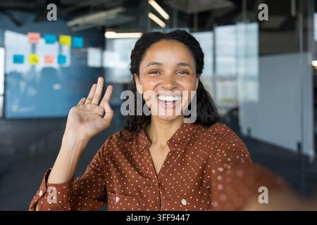 Giovane donna che sorride e saluta la telecamera, effettuando una videochiamata o salutando sui social media, comunicando allegramente da un moderno spazio di lavoro d'ufficio Foto Stock