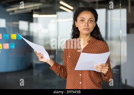 Giovane donna professionista in piedi in un ufficio contemporaneo che tiene diversi documenti cartacei, guarda la telecamera con un'espressione concentrata, rappresentando analisi e documentazione aziendale Foto Stock