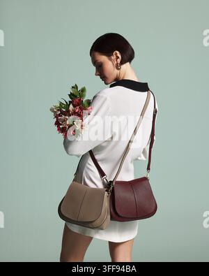 Una sofisticata giovane donna bruna in un vestito con colletto bianco tiene una borsa a tracolla in pelle marrone di alta qualità e un bellissimo mazzo di rose rosse. Foto Stock