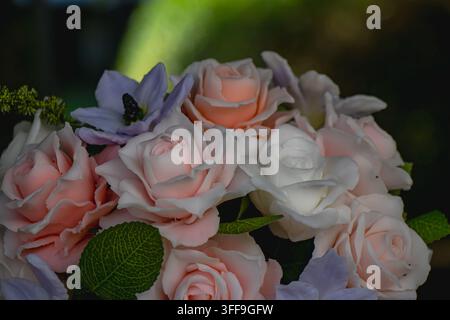 Primo piano di rose artificiali rosa confetto e bianche con fiori viola pallido e foglie verdi disposti su uno sfondo scuro per una decorazione floreale romantica Foto Stock