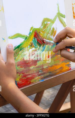 Prima vista di mani che dipingono un'opera d'arte colorata con un pennello su carta su un cavalletto di legno, mostrando un'attività artistica creativa con la texture della pittura visibile. Foto Stock