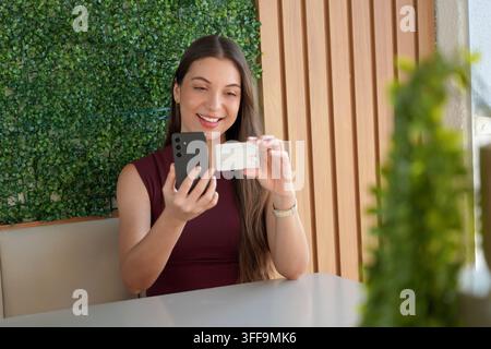 Donna casuale sorridente che tiene un telefono cellulare e legge le informazioni della carta di credito all'interno di un caffè Foto Stock