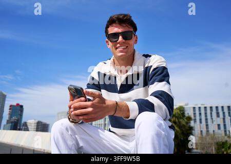 Giovane uomo che sorride mentre usa uno smartphone in un ambiente urbano Foto Stock