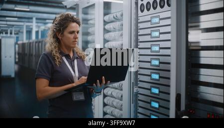 Specialista IT tiene un computer portatile per ispezionare i rack dei server e configurare l’infrastruttura cloud nella sala server del data center. La diagnostica di sistema e la manutenzione dell’hardware garantiscono un uptime di rete senza interruzioni. Foto Stock