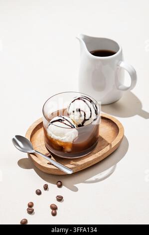 Caffè affogato con palline di gelato alla vaniglia con cioccolato ed espresso in un bicchiere su una tavola di legno su uno sfondo chiaro con chicchi di caffè, brocca Foto Stock