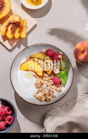 Yogurt greco con granola d'avena o muesli, pesche e lamponi in una ciotola su uno sfondo chiaro con frutta fresca, bacche e ombra mattutina. Sano an Foto Stock