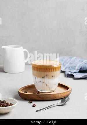 Caffè Dalgona. Caffè istantaneo montato in un bicchiere con ghiaccio su una tavola di legno su uno sfondo grigio con chicchi di caffè, cucchiaio e brocca del latte. Il concetto o Foto Stock