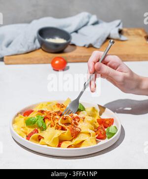 Mano che tiene una forchetta con tagliatelle o pappardelle con salsa di pomodoro, basilico e parmigiano su un piatto su sfondo chiaro. Tradizionalmente fatto in casa Foto Stock