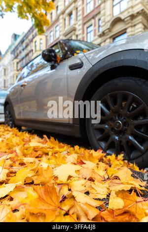 Vista panoramica del frontalino Moody, moderno parabrezza grigio per auto ricoperto di foglie cadute giallo dorato parcheggiato bagnato strada della città giorno di pioggia autunnale. Coperto Foto Stock