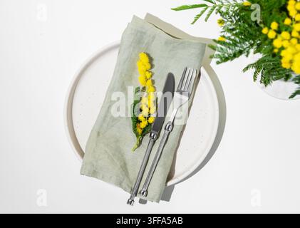 Impostazione festiva della tavola per la celebrazione primaverile della festa della donna, di un matrimonio o della festa della mamma con fiori di mimosa gialli su uno sfondo chiaro con luce solare. Res Foto Stock