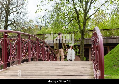 Coppia romantica che cammina sul ponte dei lucchetti dell'amore in un parco primaverile con alberi verdi e un sentiero di legno. Foto Stock
