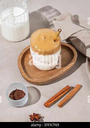 Bevanda montata coreana Dalgona coffee su un piatto di legno su uno sfondo di cemento con spezie e luce solare. Bevanda con caffè istantaneo, zucchero, latte e ghiaccio Foto Stock