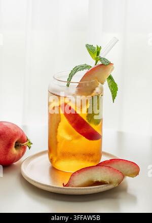 Tè freddo alla pesca con menta in un bicchiere su un tavolo bianco vicino alla finestra con frutta fresca e luce solare. Concetto di bevanda estiva rinfrescante per il menu di bar o caffè. Foto Stock
