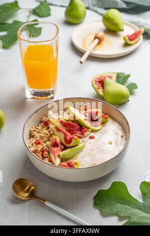 Yogur naturale con granola, fichi e miele in una ciotola su uno sfondo chiaro con un bicchiere di succo d'arancia, cucchiaio, frutta e foglie. Concetto di sano a Foto Stock