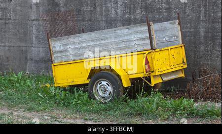 Rimorchio utilitario giallo arrugginito con pannelli laterali in legno parcheggiato sull'erba verde contro uno sfondo di muro di cemento grigio Foto Stock