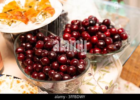 Due ciotole di vetro piene di ciliegie rosso scuro lucenti sono disposte su una tovaglia a fantasia. La frutta fresca è mostrata in primo piano, con piatti di cibo Foto Stock
