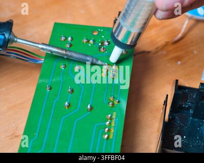 Saldatura di un circuito elettronico - Un primo piano mostra una persona che utilizza un saldatore e una pompa di saldatura per lavorare su un circuito elettronico verde Foto Stock