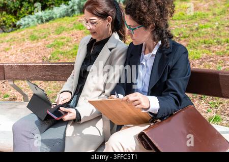 Donne d'affari che collaborano su una panchina del parco discutendo di un progetto che utilizza la tecnologia Foto Stock