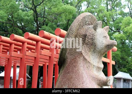Statua di pietra di una tanuki femminile in piedi su un grande piedistallo di roccia accanto a porte torii vermiglie e a una segnaletica informativa anglo-giapponese a Y Foto Stock