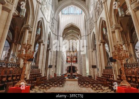 Madrid, Spagna: 7 apr. 2025 – Stalli di coro in legno ornati e alti candelabri dorati delimitano una navata di marmo a scacchi che conduce verso un crocifisso lontano e Foto Stock