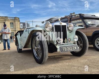 Vista frontale a basso angolo di un classico roadster MG vintage bianco parcheggiato in una mostra all'aperto con un edificio storico in pietra sullo sfondo. Foto Stock