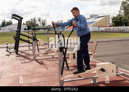 Uomo d'affari che fa esercizi in palestra. Un uomo in divisa si riscalda sui simulatori allo stadio. Esercizi sportivi dopo il lavoro d'ufficio. Il f Foto Stock