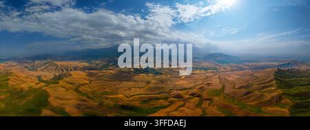 Ampia vista aerea panoramica cattura colline aspre in primo piano che si trasformano in pianure agricole e montagne lontane nel Kirghizistan. Un cielo drammatico presenta un blu brillante e nuvole sparse, con la luce del sole che illumina il paesaggio. Foto Stock