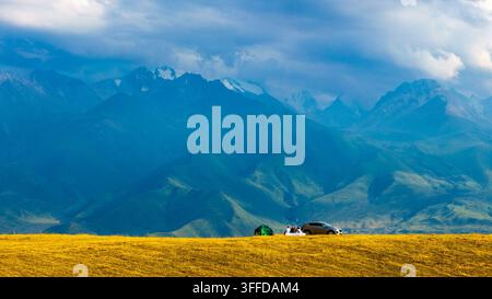 Un gruppo di campeggiatori si gode una giornata tranquilla in un vasto paesaggio con montagne blu e un campo d'erba dorata. Una tenda verde e un SUV argentato sono visibili in primo piano. Foto Stock