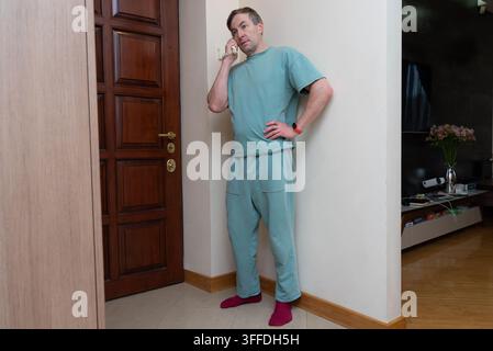 Un uomo di 40 anni in abbigliamento da salotto verde si trova in un corridoio di casa e parla al telefono fisso bianco. Foto Stock