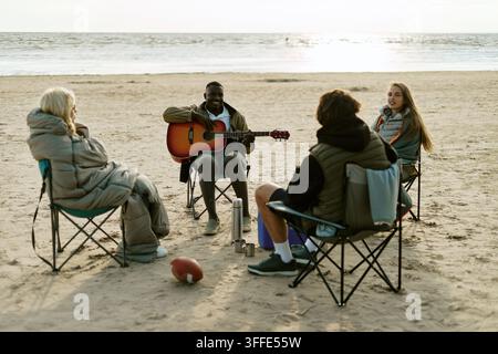 Amici diversi che campeggiano sulla spiaggia e suonano la chitarra all'aperto Foto Stock