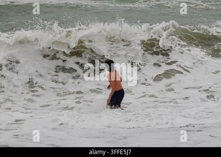 Una persona in piedi nell'acqua mentre un'onda grande si infrange su una spiaggia di ciottoli a Brighton, in Inghilterra. Foto Stock