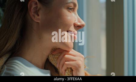 Primo piano di una donna caucasica di mezza età felice che sorride guardando fuori dalla finestra avvolta in una coperta calda, donna adulta sorride, riposa, si rilassa, pensa, allegra Foto Stock