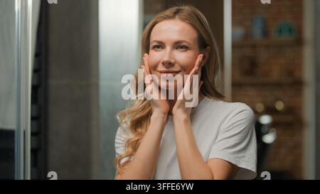 Donna caucasica attraente di mezza età che massaggia guance e collo con auto-massaggio anti-invecchiamento a casa al mattino, bella e carina donna che applica Foto Stock
