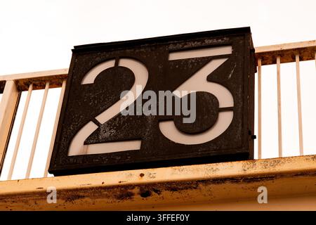 Il numero 23 è visualizzato su un cartello, composto da uno sfondo nero su un pannello quadrato attaccato a ringhiere arrugginite Foto Stock