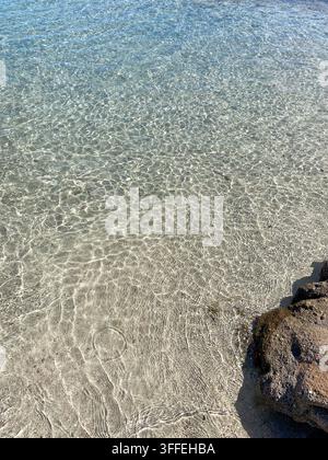 Sfondo di acqua di mare turchese cristallina con riflessi di raggi solari e caustiche sul fondale sabbioso poco profondo vicino a una roccia. Foto Stock