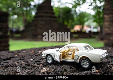 Ford Mustang 1964 1/2 modello ridotto in crema con portiera aperta, angolazione frontale accanto a una vecchia stupa nel Parco Storico di Si Satchanalai, Thailandia Foto Stock