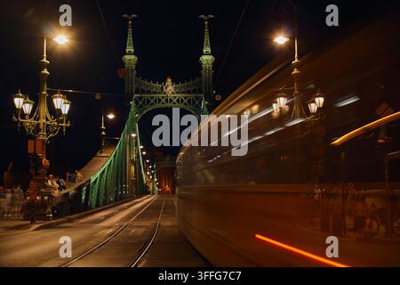 Budapest, Ungheria - 26 agosto 2026: Vista notturna a lunga esposizione di un tram pubblico che sfreccia con sfocatura del movimento oltre lampioni decorati e luminosi sull’hist Foto Stock