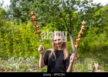 Giovane donna che tiene spiedini di carne grigliata in giardino. Giovane donna sorridente tiene due spiedini di carne grigliata all'aperto in un giardino verdeggiante durante un Foto Stock