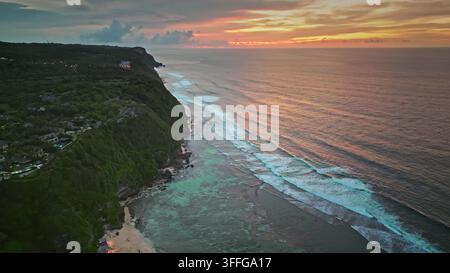 Costa tropicale della penisola di Bukit nella luce della sera, Bali, Indonesia. Scogliere verdi lussureggianti, acqua turchese tranquilla dei barriere coralline, onde oceaniche che si infrangono e cielo vivace del tramonto. Panorama di volo aereo con drone Foto Stock