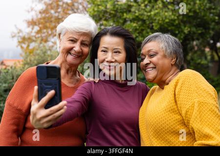 Diverse amiche che posano insieme nel parco tenendo uno smartphone e indossando caldi maglioni di lana Foto Stock