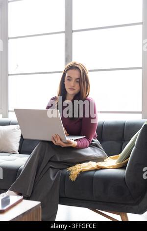 Donna seduta su un divano grigio scuro trapuntato che usa un laptop argentato in salotto vicino a uno smartphone Foto Stock