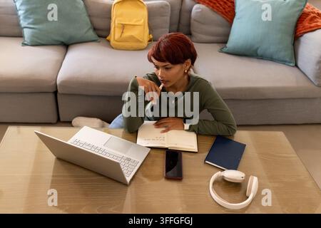 Donna afroamericana appoggiata su un tavolino basso a casa, con una penna sopra un taccuino, che usa un laptop Foto Stock