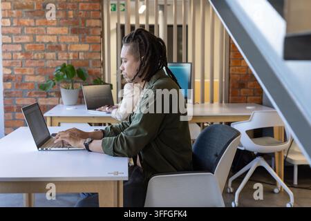 Diversi colleghi che lavorano in un ufficio condiviso, un uomo africano in camicia verde oliva che digita su un laptop Foto Stock