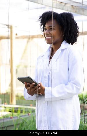 Scienziata donna di mezza età in piedi nella stanza dei bambini con un camice da laboratorio bianco, che tiene un tablet vicino a un traliccio Foto Stock